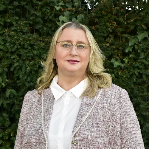 Frau mit blonden mittellangen Haaren, Brille, weißer Bluse und kariertem Blazer vor grüner Hecke.